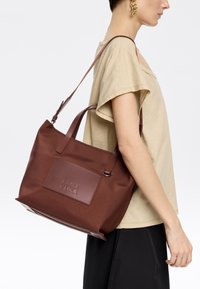 Bolsa de tela marrón con un parche rectangular de cuero que presenta el nombre de la marca en relieve. Dos asas robustas y un diseño minimalista.