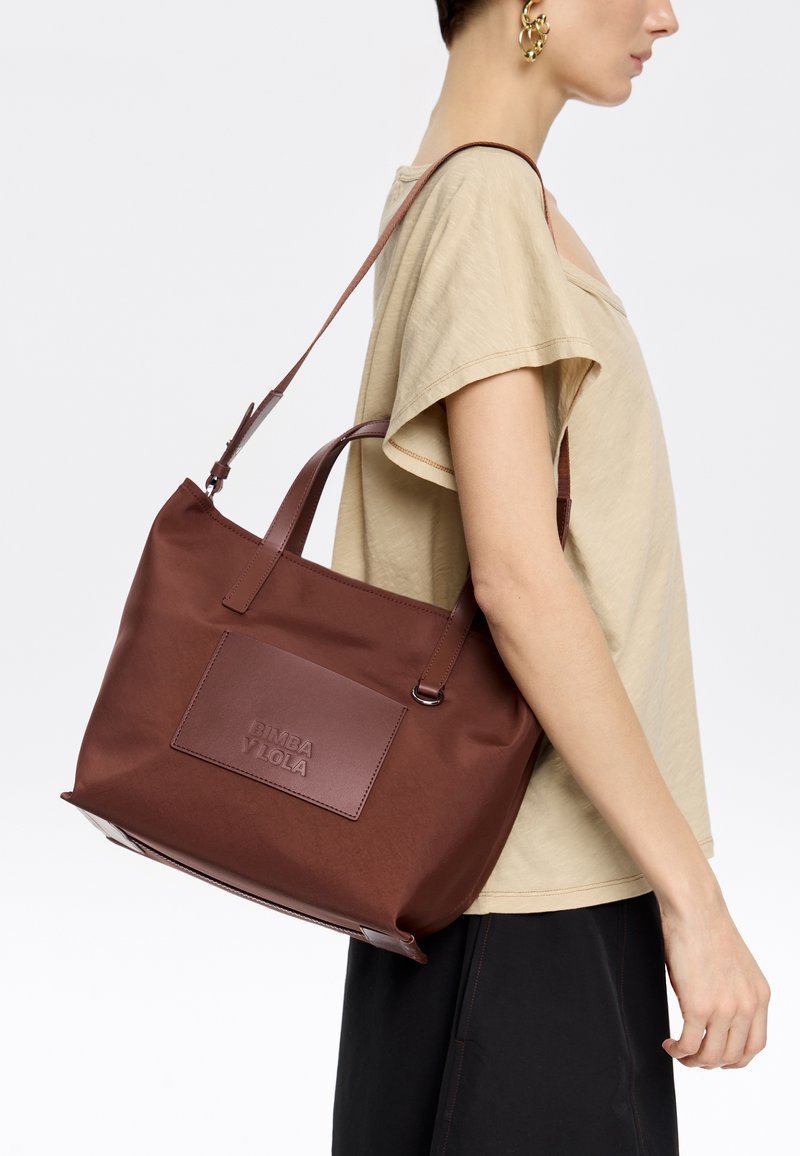 Bolsa de tela marrón con un parche rectangular de cuero que presenta el nombre de la marca en relieve. Dos asas robustas y un diseño minimalista.