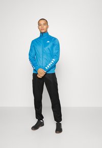 Muž stojaci so založenými rukami, oblečený v modrej zipsovej bunde s bielymi logami na rukávoch a na hrudi, čiernych nohaviciach a čiernych teniskách.