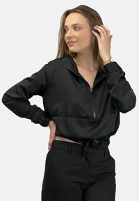 Czarna kurtka zapinana na zamek z krótkim krojem, wykonana z gładkiego materiału. Posiada elastyczne mankiety i regulację w pasie, zestawiona z czarnymi spodniami.