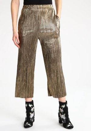 Femme portant un pantalon court plissé doré brillant et des bottines à talons noires brodées de fleurs, debout devant un fond blanc.