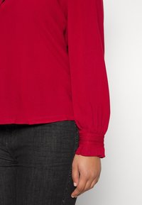 Personne portant un chemisier rouge à manches longues avec des poignets froncés et un pantalon foncé ajusté avec un détail de rayure latérale subtile, debout sur un fond blanc.