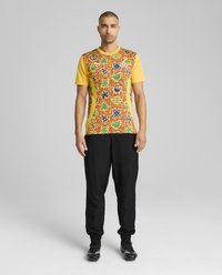 Gul atletisk skjorte med et mønstret forsiden med geometriske designs i grøn, blå og rød; parret med sorte joggerbukser og sort fodtøj.