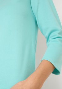 Camiseta de manga larga en color turquesa claro con una textura suave. Presenta un escote redondeado y puños que llegan justo por encima del codo.