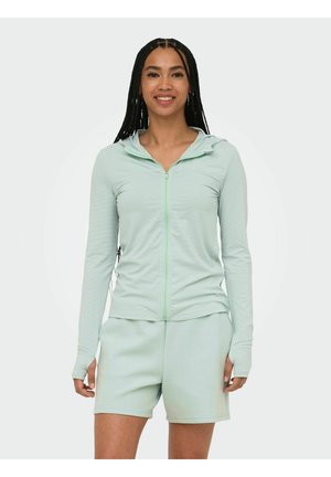 Naine pika punutud soenguga, naeratab, seljas heleroheline lukuga dressipluus ja sobivad lühikesed püksid, monotoonse taustaga.