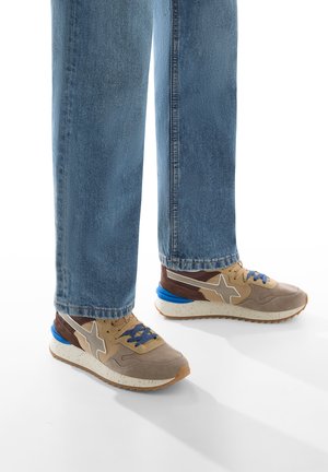 Le sneakers presentano una tomaia in suede beige con accenti marroni, un contrafforte blu e una suola intermedia bianca texture. Abbinati a jeans in denim azzurro chiaro.