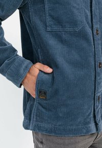 Blå kordfløjsjakke med knaplukning. Har en brystlomme og et logo-patch nær kanten. Materialet har en vertikal tekstur.