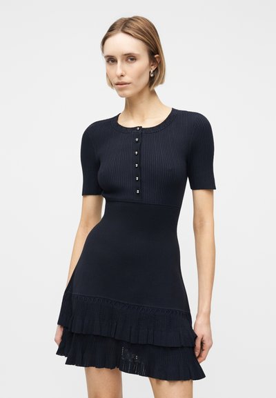 Γυναίκα με κοντά ξανθά μαλλιά που φοράει στενή μαύρη φόρεμα με κοντά μανίκια, ριπέν κουμπωτό τοπ και φούστα με στρώσεις βολάν.