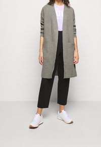 Kvinna iklädd en grå lång kofta, lila T-shirt, svarta korta byxor och vita sneakers med gummisula, stående mot en plain bakgrund.