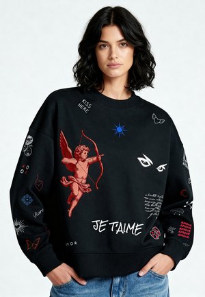 Jeune femme portant un sweat-shirt noir avec un dessin de chérubin rouge, le texte blanc "JE T'AIME" et divers petits symboles et mots colorés.