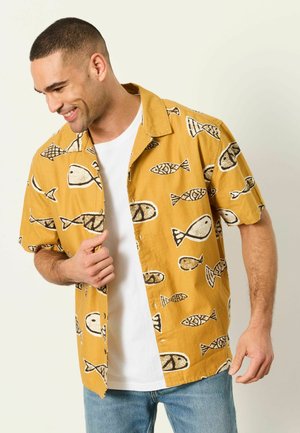 Mann lächelt und schaut nach unten, trägt ein senfgelbes Kurzarmhemd mit Fischmotiven über einem weißen T-Shirt und blauer Jeans.