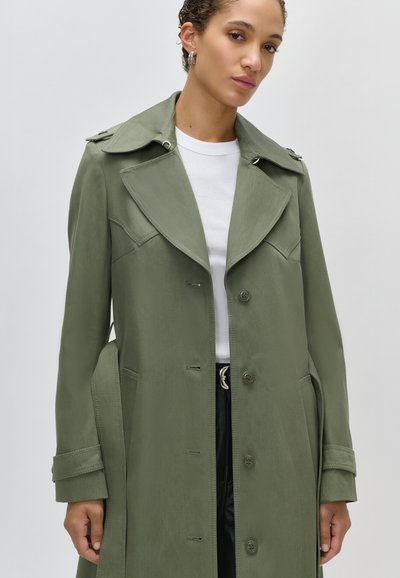 Γυναίκα που φοράει ένα χακί καμπαρντίνα πάνω από ένα λευκό πουκάμισο, στέκεται μπροστά σε ένα μονόχρωμο ανοιχτό γκρι φόντο.