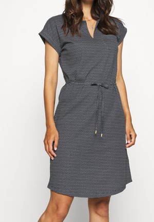 Femme portant une robe noire à manches courtes avec des pois blancs, un col en V et une ceinture à la taille, debout devant un fond uni.