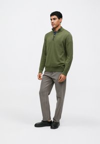 Παρντό μπλούζα πλεκτή σε ελιά πράσινο με φερμουάρ στο τέταρτο, στρογγυλό γιακά και μακριά μανίκια, συνδυασμένη με γκρι παντελόνια σε χαλαρή γραμμή και μαύρα παπούτσια.