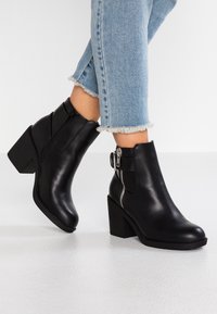 Des bottines noires en cuir avec un talon épais, une fermeture éclair sur le côté et des accents texturés, assorties à un jean en denim avec ourlet effiloché.