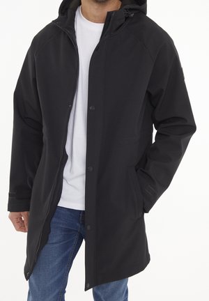 Mand iført en sort hættefrakke med lynlås og knapper foran over en hvid t-shirt og blå jeans, med den ene hånd i frakkens lomme.