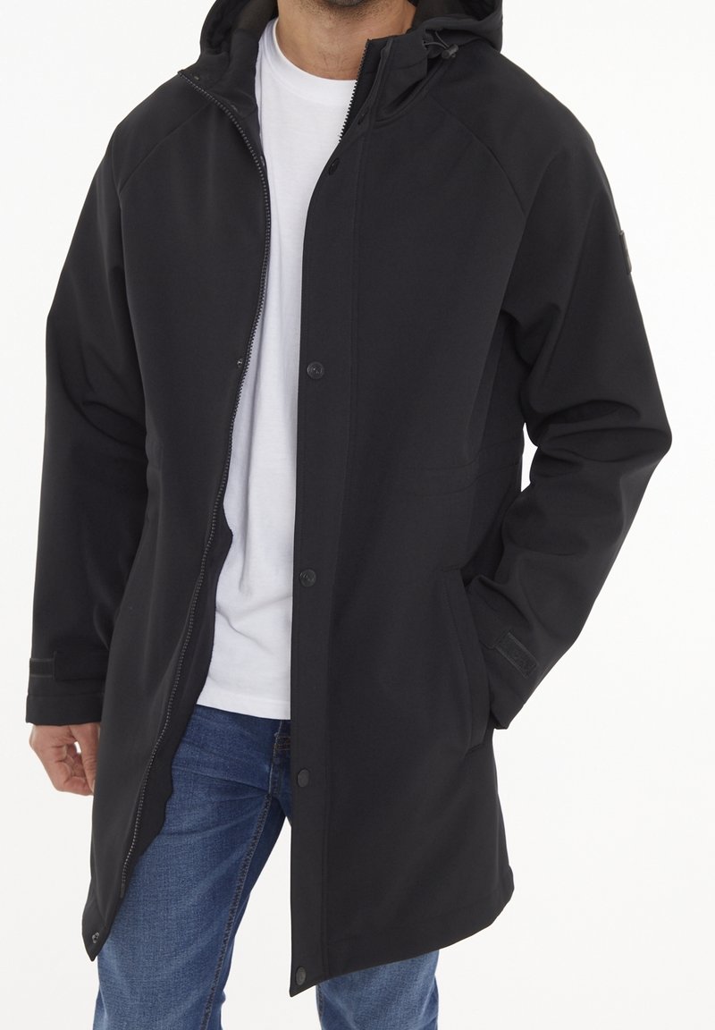 Homme portant un manteau noir à capuche avec fermeture éclair et boutons à l'avant, par-dessus un t-shirt blanc et un jean bleu, une main dans la poche du manteau.