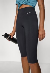 Shorts de vélo noirs ajustés avec une taille haute, fabriqués en matière extensible, arborant un petit logo Nike blanc à l'avant. Tient une bouteille d'eau noire.