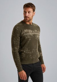 Groene gebreide trui met een textuurpatroon en een vervaagd effect, met de tekst "ME LEGEND" op de borst. Geribde manchetten en zoom.