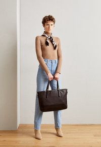 Suéter marrón de canalé, jeans de tiro alto azul claro y botas de tobillo beige. Sosteniendo un gran bolso tote de cuero negro con dos asas.