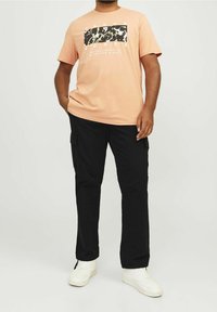 Jasnoróżowa koszulka z czarno-białym nadrukiem graficznym; czarne spodnie typu cargo; białe trampki. Klasyczny okrągły dekolt i regularny krój.