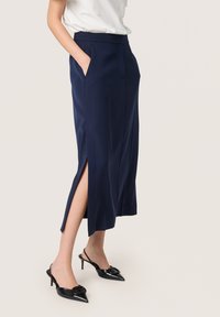 Marineblaue Midirock mit zwei seitlichen Taschen, ausgeprägten Seitenschlitzen und einem glatten Stoff. Kombiniert mit schwarzen, spitzen Slingbacks.