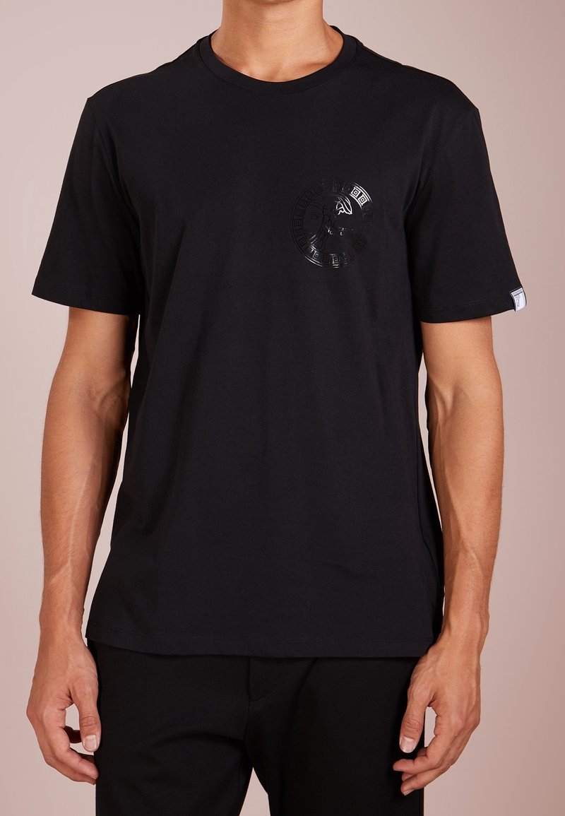 Homme portant un t-shirt noir uni à manches courtes avec un motif circulaire discret et brillant sur le côté gauche de la poitrine, associé à un pantalon noir.