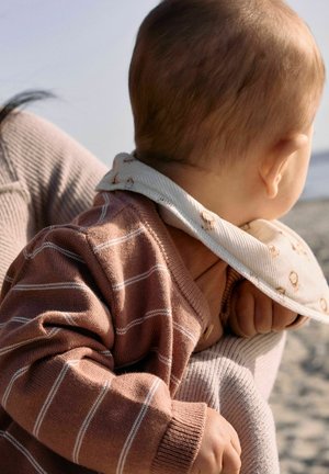 Baby trägt braun gestreiften Pullover und weißes Lätzchen, gehalten von einem Erwachsenen mit beiger, gerippter Kleidung, im Hintergrund ein sandiger Strand und Himmel.
