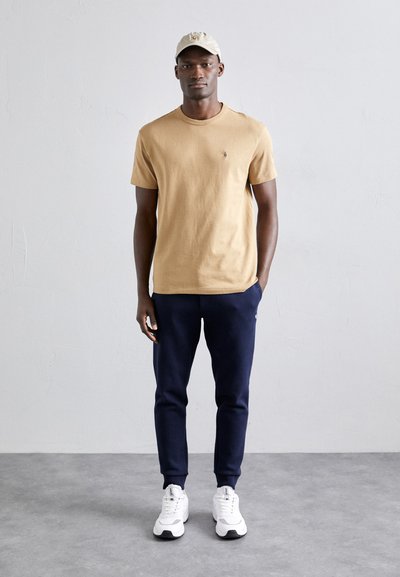 Beige cotton t-shirt with a small logo, navy fleece pants, and white sneakers. The model wears a light beige cap, standing against a gray backdrop.