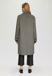 Grijze wollen overjas met een ingesneden kraag, enkele split aan de achterzijde en met knopen afgewerkte manchetten. Gepaard met zwarte, cropped broek en schoenen.