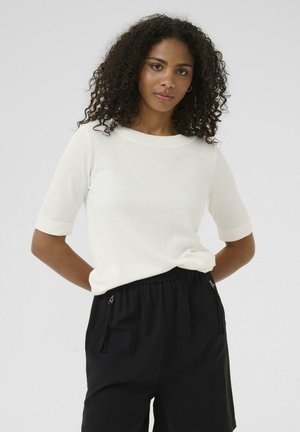 Weißes kurzärmliges Oberteil mit Rundhalsausschnitt, das eine weiche Textur aufweist. Kombiniert mit schwarzen Shorts, die über seitliche Reißverschlusstaschen verfügen.