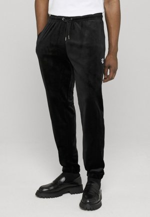Homme portant un pantalon de jogging en velours noir avec des poignets élastiques, associé à des mocassins noirs épais et une chemise blanche, debout devant un fond uni.