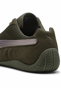 Zapatilla deportiva en verde olivo con parte superior de malla, acentos de ante, suela de goma texturizada y una franja morada en el lateral.