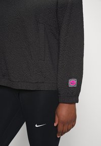 Zwarte textuur trui met een zak, met een roze Nike logo patch op de mouw. Gecombineerd met zwarte leggings versierd met een witte Nike swoosh.