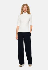 Weißes Turtleneck-Oberteil mit kurzen Ärmeln, kombiniert mit lockeren schwarzen Hosen und hellen Sneakern. Glatte Textur, taillierte Passform.