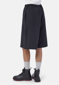 Persona che indossa culottes nere fino al ginocchio, calze grigie e stivali neri con lacci e suole rosse, in piedi contro uno sfondo bianco.
