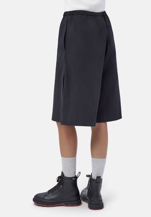 Persona che indossa culottes nere fino al ginocchio, calze grigie e stivali neri con lacci e suole rosse, in piedi contro uno sfondo bianco.