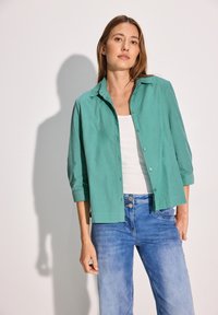 Camisa de botones turquesa con mangas arremangadas, combinada con jeans de un azul claro. La tela parece liviana y tiene una textura suave.