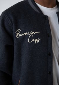 Dark blue buttoned jacket with white cursive "Bavarian Caps" embroidered on the chest, worn over a plain white T-shirt.