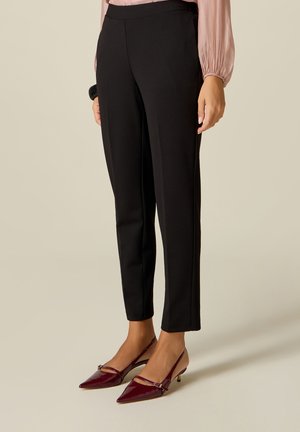 Femme portant un pantalon noir slim, un chemisier rose et des talons slingback bordeaux à bout pointu, debout devant un fond beige.