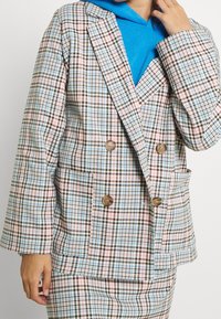 Tuplavideinen bleiseri ruutukuosisena, joka sisältää sinisiä, vaaleanpunaisia ja ruskeita sävyjä. Mukana on kaksi taskua ja puunapit.