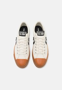 Baskets en toile blanche avec embout en caoutchouc marron et semelle beige, présentant le logo noir "Converse All Star" sur la languette et le côté.