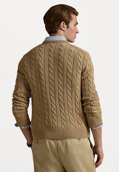 Kabelgebreide trui in een beige kleur, met textuurpatronen, geribde manchetten en zoom, gedragen over een lichtblauw overhemd met kraag.