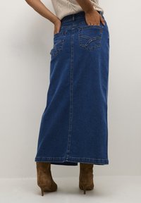 Falda maxi de mezclilla en azul oscuro, con bolsillos traseros, corte recto y costura central. Combinada con botas de tacón alto de ante color marrón.