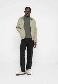 Άνδρας που στέκεται μπροστά σε λευκό τοίχο, φοράει μπεζ σακάκι, γκρι πουλόβερ, μαύρο παντελόνι και εκρού αθλητικά παπούτσια με καφέ σόλες.