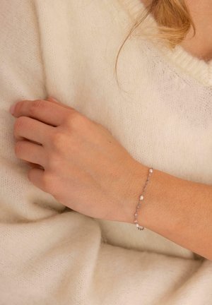 Feines Silberarmband mit kleinen weißen Perlen, getragen am linken Handgelenk über einem cremefarbenen Pullover.