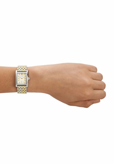 Muñeca con reloj de metal bicolor plateado y dorado con esfera rectangular, de color claro, y marcadores y agujas de hora dorados.