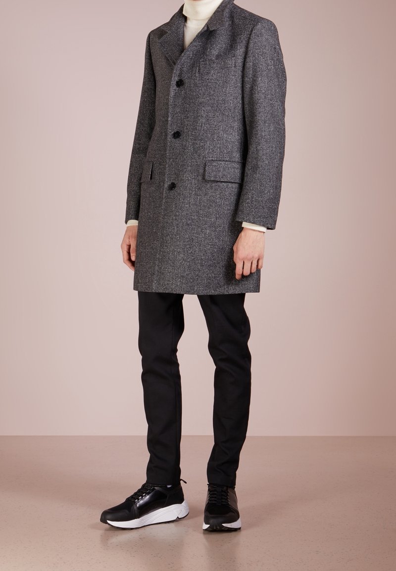 Grå tweed overfrakke med brede klapper, enkeltknap, to frontlommer, båret over en elfenbensfarvet turtleneck, parret med sorte bukser og sneakers.