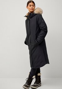 Zwarte parka met nepbont capuchon, twee voorkezen en een rits. Gekoppeld aan zwarte jeans en bruine waterdichte laarzen met bontafwerking.