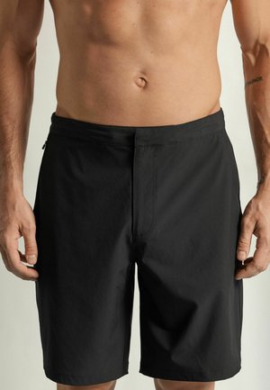 Mand ifører almindelige sorte knælange shorts, står med armene afslappet ned langs siderne, viser bar overkrop og tatovering på højre underarm.